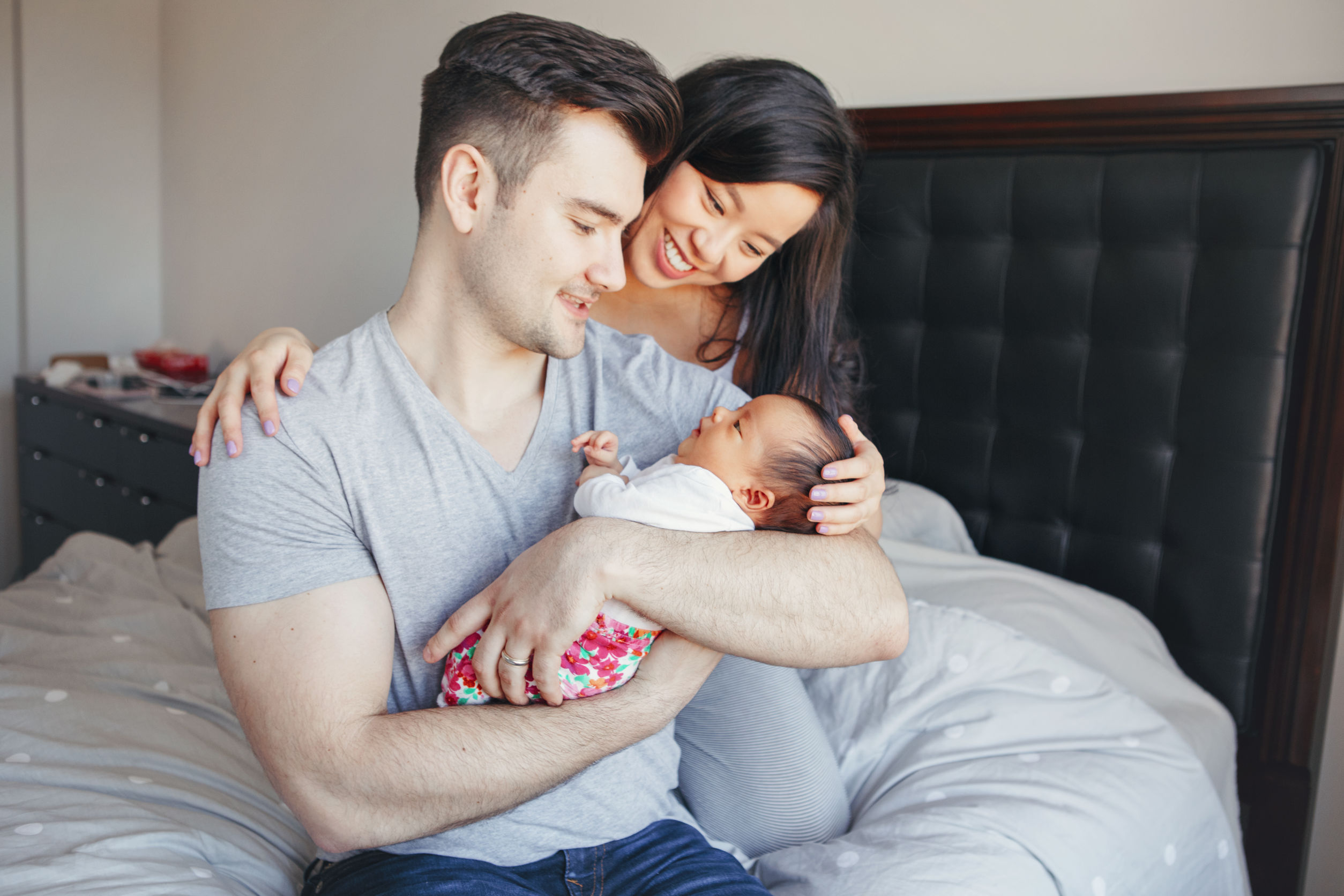 Portrait of beautiful smiling Chinese Asian mother and Caucasian father with mixed race newborn infant baby son daughter. Happy family in bedroom. Home lifestyle authentic natural moment.